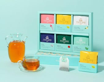 May include: A teal tea gift set with six boxes of tea bags, a glass teapot, and a teacup with a saucer. The tea bags are in various colors, including pink, yellow, and green, with the brand name "TICLIPS" on each box.