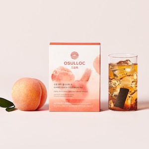 May include: A still-life image featuring a ripe peach, a box of "OSULLOC Sunset Peach Cold Brew Tea," and a glass of iced tea. The box has peach illustrations and Korean text. The tea appears amber-colored.