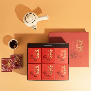 Könnte beinhalten: Draufsicht auf ein Teeservice mit einer weißen Keramik-Teekanne, einer kleinen Tasse Tee und einer Schachtel mit Teebeuteln. Die Teebeutel und -schachteln sind rot mit goldfarbenen Buchstaben. Der Hintergrund ist warm und neutral.