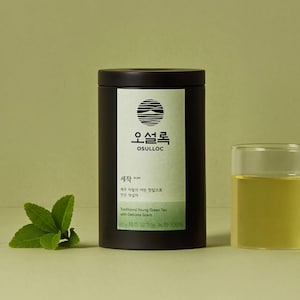 Può includere: Una lattina di tè cilindrica nera con un'etichetta bianca con testo coreano e un disegno circolare. L'etichetta recita "OSULLOC" e "Traditional Young Green Tea with Delicate Scent". Sono visibili anche un bicchiere di tè verde e foglie di tè.