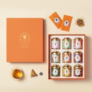 May include: An orange gift box set of tea tins with gold lids. The box contains nine different tea flavors in small, decorative tins. Two tea bags and a cup of tea are also visible. The box is labeled "SSANGGYE TEA GOLD JUBILEE COLLECTION."