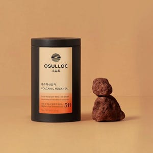 Könnte beinhalten: Ein schwarzer zylindrischer Behälter mit Osulloc Volcanic Rock Tea mit einem weißen Etikett und dem Text "OSULLOC" und "VOLCANIC ROCK TEA". Rechts vom Behälter befindet sich eine kleine, dunkelbraune Felsformation. Der Behälter hat ein Nettogewicht von 176g.
