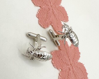 Puede incluir: Un par de gemelos de plata con un diseño de insecto detallado. Los gemelos se muestran sobre un fondo de tela rosa.