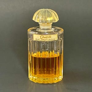 Puede incluir: Un frasco de perfume de vidrio transparente con lados estriados verticales que contiene líquido ámbar. El frasco presenta una etiqueta con el texto Quadrille BALENCIAGA y está rematado con un tapón translúcido, acanalado y en forma de cúpula.