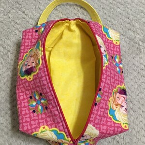 May include: Pink zippered pouch with a yellow handle and a yellow lining. The pouch features a repeating pattern of pink and white teardrop shapes and a floral design. The pouch has a print of two girls with blonde hair.