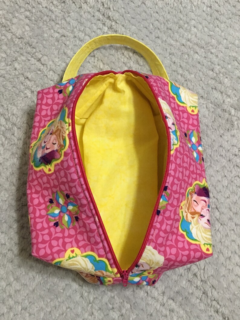 May include: Pink zippered pouch with a yellow handle and a yellow lining. The pouch features a repeating pattern of pink and white teardrop shapes and a floral design. The pouch has a print of two girls with blonde hair.