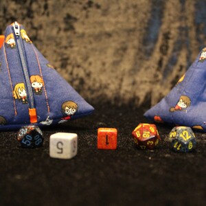 May include: Two blue fabric dice bags with a Harry Potter character print. The bags are triangular and have orange zippers. There are several dice in front of the bags, including a red d20, a blue d12, a white d6, a red d4, a blue d20, and a yellow d4.