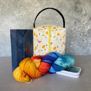 May include: A white fabric tote bag with a bee and honeycomb pattern. The bag has a zipper closure and a black handle. The bag is sitting on a table with a blue leather wallet and a colorful yarn skein.