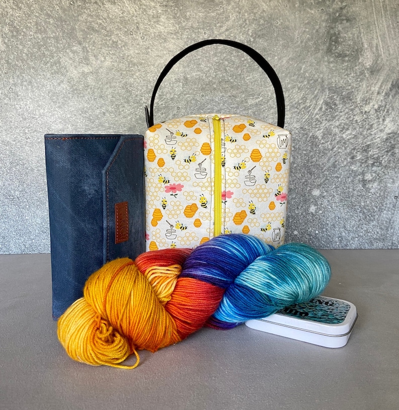 May include: A white fabric tote bag with a bee and honeycomb pattern. The bag has a zipper closure and a black handle. The bag is sitting on a table with a blue leather wallet and a colorful yarn skein.