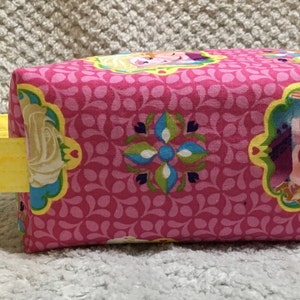 May include: Pink fabric tote bag with a yellow handle. The bag features a repeating pattern of pink and green leaves and a floral design. The front of the bag has a picture of two girls with blonde hair, one with a blue dress and the other with a purple dress.