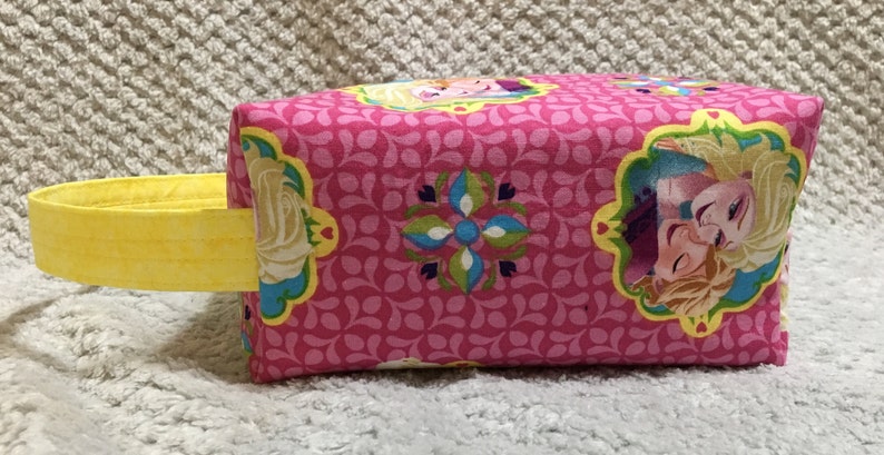May include: Pink fabric tote bag with a yellow handle. The bag features a repeating pattern of pink and green leaves and a floral design. The front of the bag has a picture of two girls with blonde hair, one with a blue dress and the other with a purple dress.