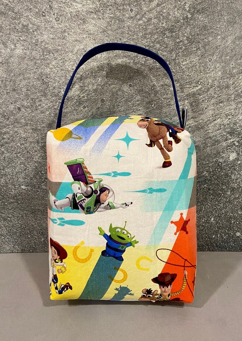 May include: A colorful fabric doorstop with a blue handle. The fabric features a pattern of characters from the Toy Story movie franchise, including Woody, Buzz Lightyear, and the alien.