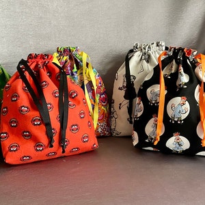 May include: Six colorful drawstring bags with various cartoon character prints. The bags are made of fabric and have black drawstrings. The bags are arranged in a row on a gray surface.