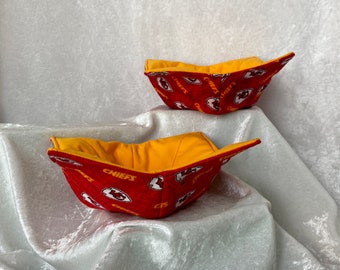 May include: Two red and yellow fabric bowls with a Kansas City Chiefs logo pattern. The bowls are folded and sitting on a white surface.