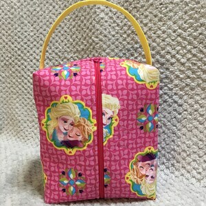 May include: A pink fabric zippered pouch with a yellow handle. The pouch features a repeating pattern of pink leaves and flowers. The front of the pouch has a picture of two girls with blonde hair, one hugging the other. The girls are wearing blue and white dresses.