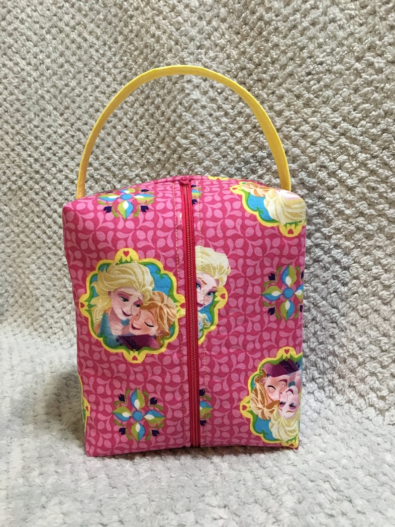May include: A pink fabric zippered pouch with a yellow handle. The pouch features a repeating pattern of pink leaves and flowers. The front of the pouch has a picture of two girls with blonde hair, one hugging the other. The girls are wearing blue and white dresses.
