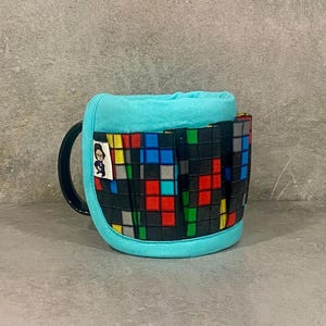 Peut inclure: Un manchon de tasse bleu turquoise avec un motif géométrique noir, rouge, bleu et jaune. Le manchon a une petite étiquette avec une image de dessin animé d'une personne portant des lunettes.