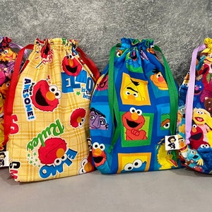 May include: Four colorful drawstring bags featuring Sesame Street characters. The bags are made of fabric and have different patterns. The bags are lined up in a row on a gray background.