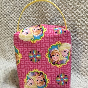 May include: A pink fabric tote bag with a yellow handle. The bag features a repeating pattern of pink and green floral designs with images of two girls from the movie Frozen.