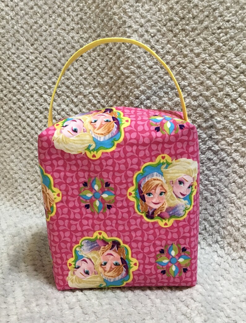 May include: A pink fabric tote bag with a yellow handle. The bag features a repeating pattern of pink and green floral designs with images of two girls from the movie Frozen.