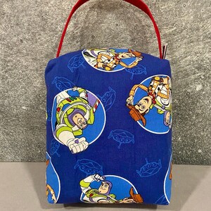 May include: A blue fabric tote bag with a red handle. The bag features a repeating pattern of Toy Story characters, including Woody and Buzz Lightyear.