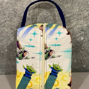 May include: A white zippered pouch with a blue handle. The pouch features a colorful print of characters from the Toy Story movie franchise, including Woody, Buzz Lightyear, and the alien characters.