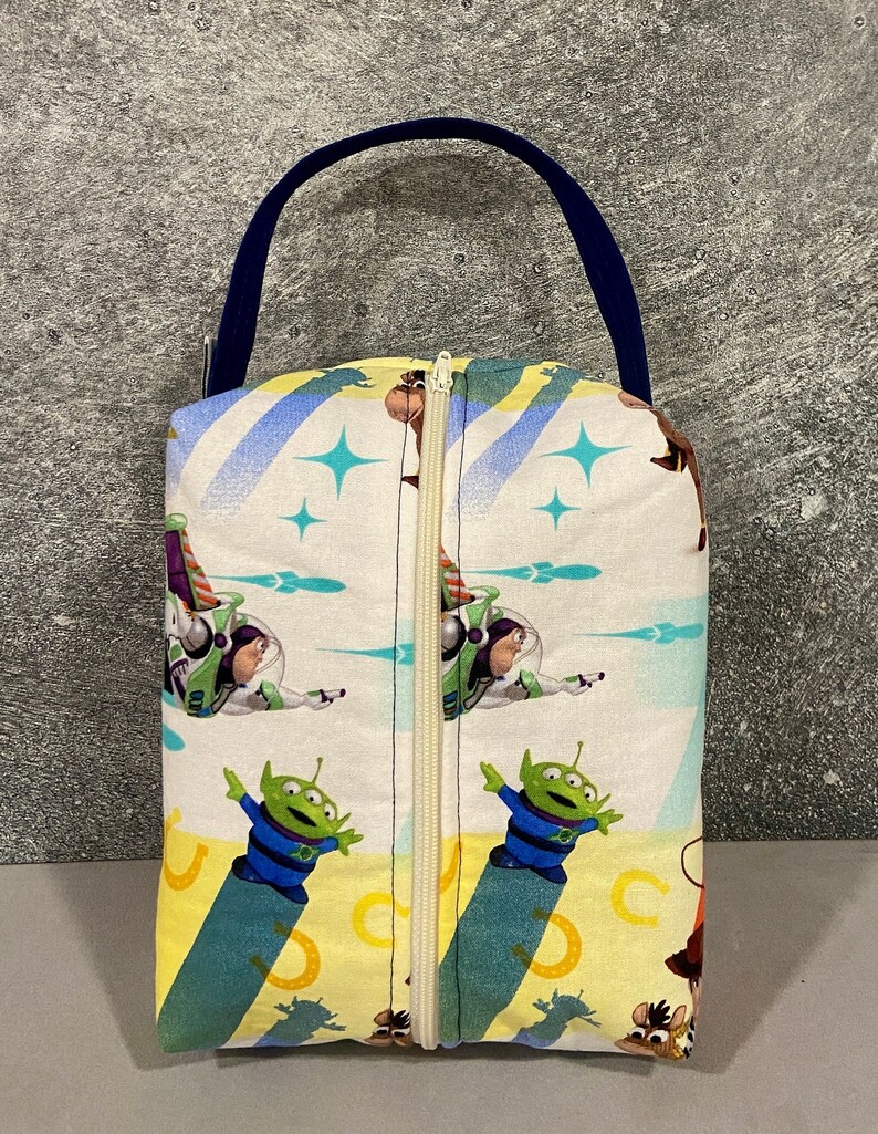 May include: A white zippered pouch with a blue handle. The pouch features a colorful print of characters from the Toy Story movie franchise, including Woody, Buzz Lightyear, and the alien characters.