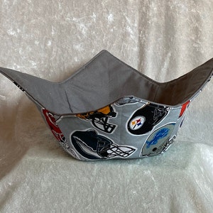 May include: A grey fabric bowl with a football helmet pattern. The bowl is made of fabric and has a grey lining. The bowl is designed to hold snacks or other small items.