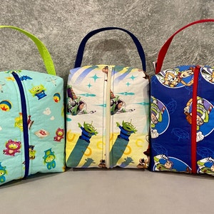 May include: Three zippered pouches with different Toy Story character prints. The pouches are blue, yellow, and green with a variety of characters including Woody, Buzz Lightyear, and the aliens. Each pouch has a zipper closure and a fabric handle.