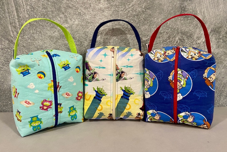 May include: Three zippered pouches with different Toy Story character prints. The pouches are blue, yellow, and green with a variety of characters including Woody, Buzz Lightyear, and the aliens. Each pouch has a zipper closure and a fabric handle.