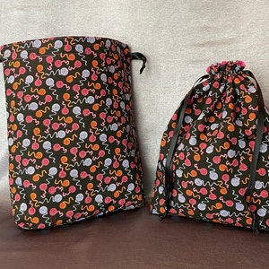 May include: Two drawstring bags with black fabric and a black and white yarn ball pattern. The larger bag has a black drawstring and the smaller bag has a pink drawstring.