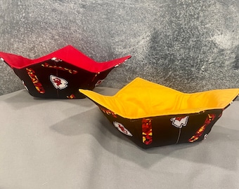 May include: Two fabric bowl cozies, one red and one yellow, with a black fabric interior featuring a repeated pattern of a football team logo and the team's name.