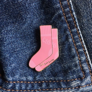 May include: A pink enamel pin in the shape of two socks, with the letters "CSS" and "S". The pin has gold edging and the words "Cozy Sock Society" are written at the bottom. The pin is set against a denim background.