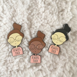 May include: Three enamel pins featuring cartoon illustrations of people with glasses and the text "Always Tired". The pins are on a white fluffy surface.
