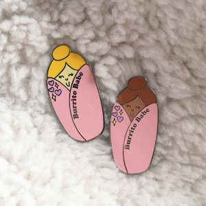 May include: Two pink and yellow enamel pins shaped like burritos with smiling faces and the text "Burrito Babe".