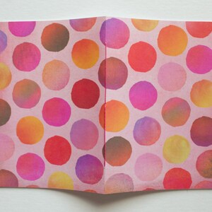 May include: Open notebook with a pink cover featuring a pattern of colorful polka dots in shades of red, pink, orange, yellow, and purple. The dots are arranged in a repeating pattern across the pages.