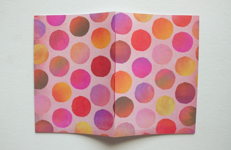 May include: Open notebook with a pink cover featuring a pattern of colorful polka dots in shades of red, pink, orange, yellow, and purple. The dots are arranged in a repeating pattern across the pages.