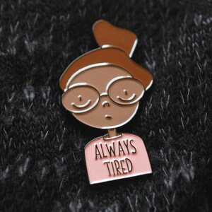 May include: A brown and pink enamel pin featuring a cartoon illustration of a person with brown hair, glasses, and a pink shirt that says "ALWAYS TIRED".