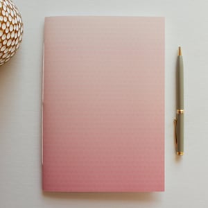 May include: A pink ombre notebook with a gray pen on a white surface.