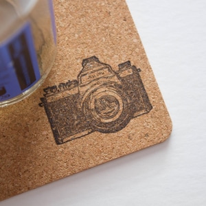 May include: A cork coaster with a dark brown vintage camera design. The coaster is square with rounded corners. A clear glass bottle with a blue label is partially visible in the upper left corner.