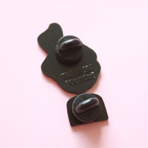 May include: Black enamel pin featuring a cartoon image of a person holding a microphone. The pin has two metal clasps on the back and the text "The Neck Don't Truss" printed on the back.