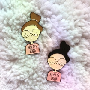 May include: Two enamel pins featuring cartoon illustrations of people with glasses and buns. Both pins have a pink shirt with the text "Always Tired" on them.