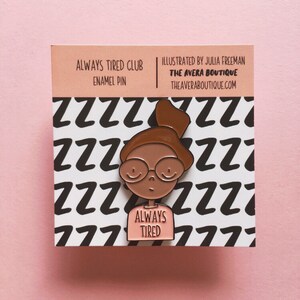 May include: A pink enamel pin featuring a cartoon illustration of a person with brown skin wearing glasses and a t-shirt that says "Always Tired". The background is a white card with a black and white pattern of the letter "Z".