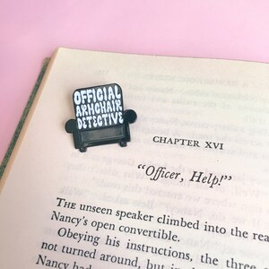 Peut inclure: &Eacute;pinglette en &eacute;mail noir en forme de fauteuil avec le texte "OFFICIAL ARMCHAIR DETECTIVE" en lettres blanches.