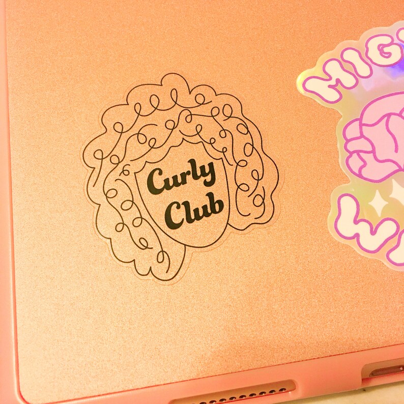May include: A pink laptop with a black and white sticker of a curly haired person with the text "Curly Club" on it. There is also a holographic sticker with the text "High" on it.