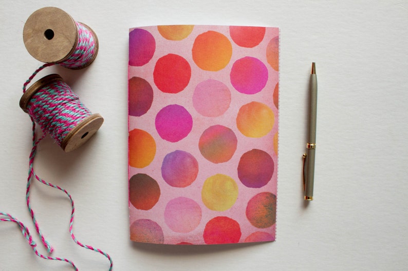 May include: A notebook with a pink cover featuring colorful watercolor circles in shades of red, orange, yellow, and purple. Two wooden spools of pink and teal twine and a silver pen are also present.