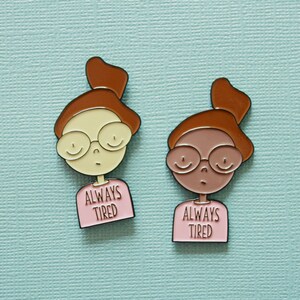 May include: Two enamel pins featuring cartoon illustrations of people wearing glasses and pink shirts with the text "Always Tired".