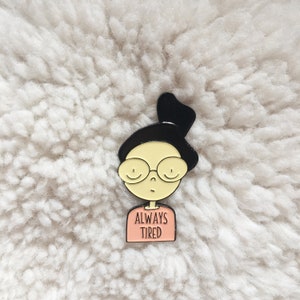 May include: Black and white enamel pin of a cartoon person wearing glasses and a shirt that says "Always Tired".
