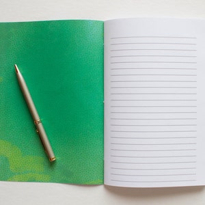 Puede incluir: Un cuaderno verde con un bol&iacute;grafo sobre la portada. El cuaderno tiene un patr&oacute;n moteado y el bol&iacute;grafo es plateado con la parte superior dorada.