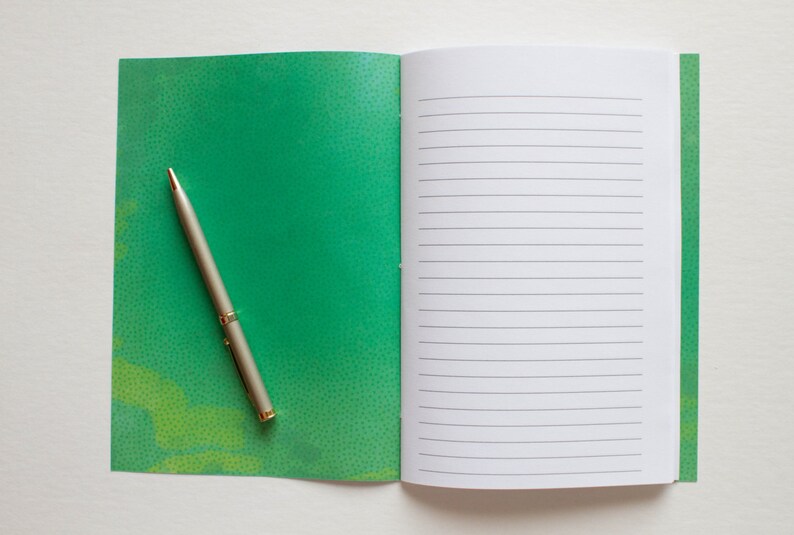 Puede incluir: Un cuaderno verde con un bol&iacute;grafo sobre la portada. El cuaderno tiene un patr&oacute;n moteado y el bol&iacute;grafo es plateado con la parte superior dorada.